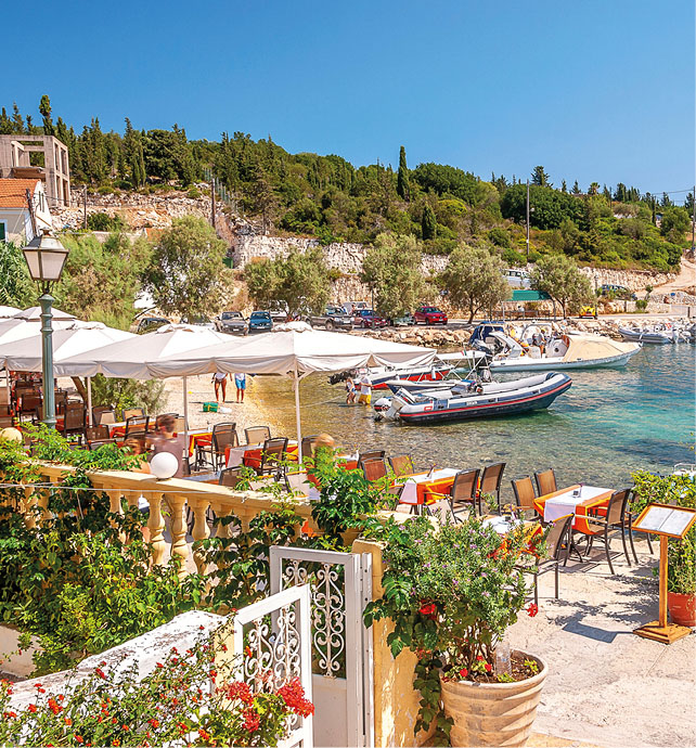 restaurant by the water with white walls, green plants with red flowers, tables outside and boats in the water 