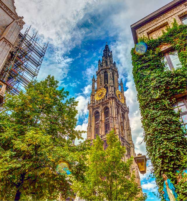 Tall spire of a cathedral behind green trees with a building covered in green vines