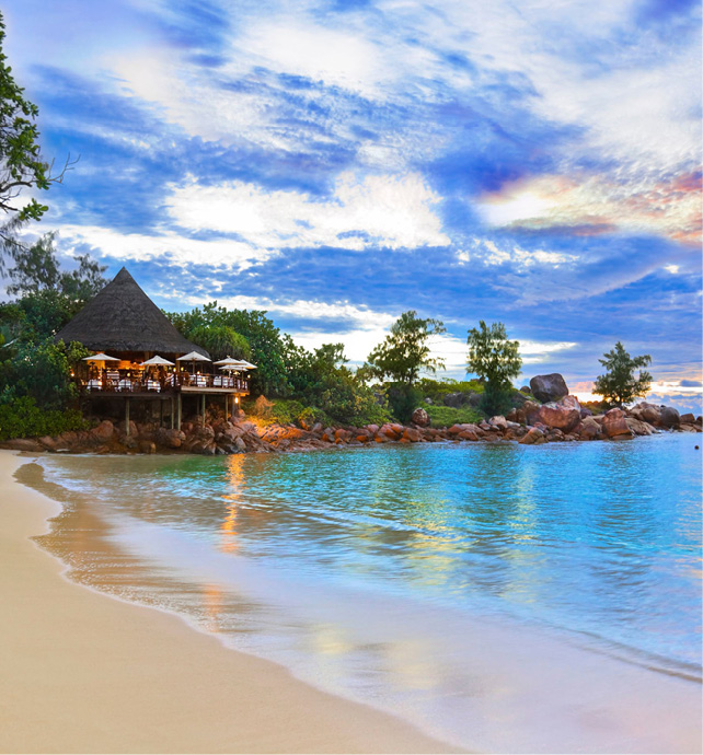 island thatched roof hut on the edge of a sandy beachfront with crystal blue water at dust with a sunset in the sky