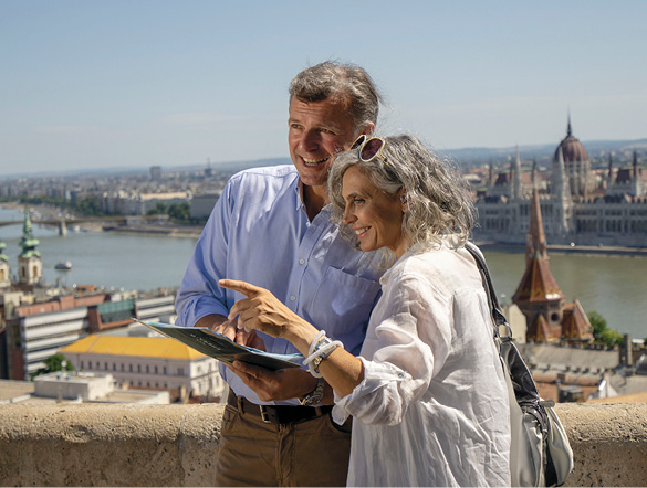 A couple reading a leaflet and smiling atop of Buda Hill overlooking the Budapest Parliament Building and Danube River