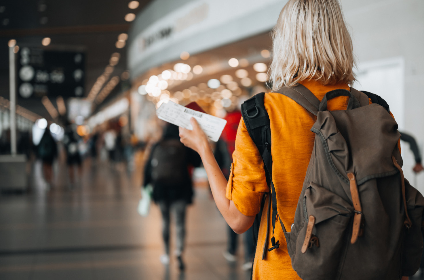 A woman in an airport carries a rucksack on her back and a plane ticket in her hand